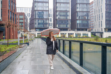 Strolling woman carries umbrella on rainy city street. Pedestrian walks along wet sidewalk near offices. Commuting travel, rain day, urban lifestyle, waterproof shelter, weather protection. Transit.