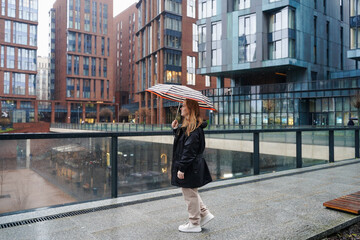 Walking woman holds umbrella on rainy city walkway. Commuter crosses wet pavement in urban district. Street travel, rain protection, waterproof canopy, autumn weather, outdoor lifestyle. Daily commute