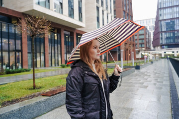 Standing woman holds striped umbrella in rain. Looking away near wet city promenade. Lifestyle commuting, bad weather, travel protection, waterproof canopy, outdoor portrait, urban street. Drizzle.