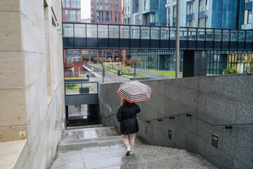 Descending woman with umbrella walks down concrete stairs in rain. Entering underpass with wet steps. Urban commute, pedestrian travel, city transport, rain protection, waterproof canopy. Mobility.