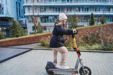 Riding electric scooter on rainy city sidewalk. Balancing micromobility transport during commute rush hour. Wet pavement reflects bad weather drizzle. Urban travel continues with scooter motion.
