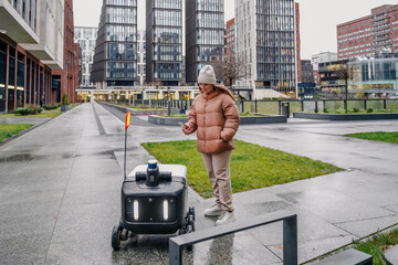 Robotic courier travels along rainy street. Customer stands with smartphone for delivery tracking, status. Autonomous logistics service provides contactless parcel transport on wet pavement.