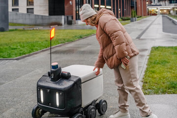Pickup process begins at autonomous delivery robot. Customer reaches to compartment while checking smartphone app, access code. Logistics enables contactless parcel handoff on rainy pavement.