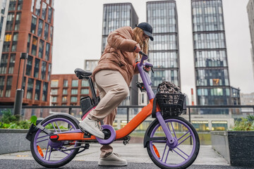 Mounting rented electric bicycle for urban commute on rainy day. Balancing pedals, handlebars before ride start. Ebike rental sharing supports last mile travel, sustainable transport, wet pavement.