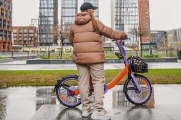 Waiting with rented electric bicycle after rainy ride on wet pavement. Holding handlebars, checking route on smartphone. Ebike rental sharing supports commute travel, sustainable transport, fitness.