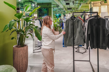 Browsing clothing rack in fashion store, customer chooses jacket for purchase. Shopping in boutique supports retail sales, wardrobe update, apparel selection, consumer lifestyle today indoors.