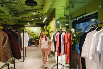 Entering fashion store, shopper walks between clothing racks. Retail boutique shopping for apparel, casual outfits. Consumer wardrobe update, purchase planning, indoor commerce, trendy collection