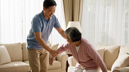 Caring young man assists his smiling elderly mother with standing up from a comfortable armchair in a bright and cozy living room setting