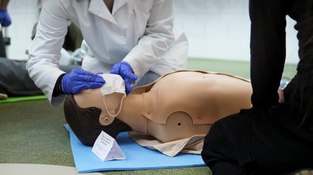 cpr training on adult mannequin with pocket mask, white coat instructor wearing blue gloves applies mask seal and tests airway, students observe on mat, stepbystep resuscitation technique,