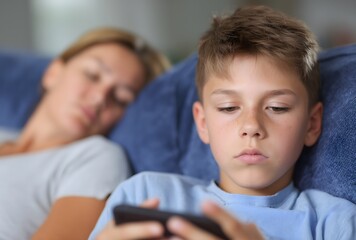 Young boy using phone on couch while mother sleeps