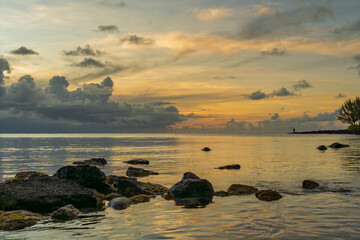 Golden sunset over calm ocean with dramatic clouds and rocky shoreline creating peaceful tropical seascape panorama