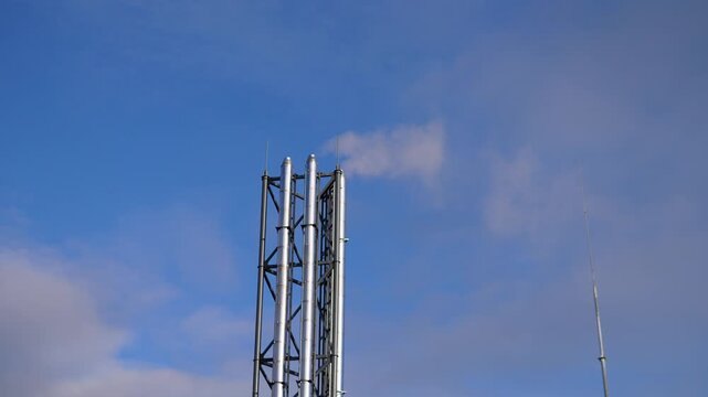 metal communication tower against blue sky, vertical lattice mast with panel antennas and slim whip, soft scattered clouds, bright morning light, highcontrast industrial detail, steady pan