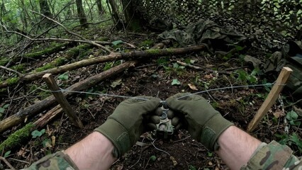Soldier in camouflage gloves setting tripwire grenade trap amid woodland forest. Military survival training with wooden stakes and netting in mossy terrain.