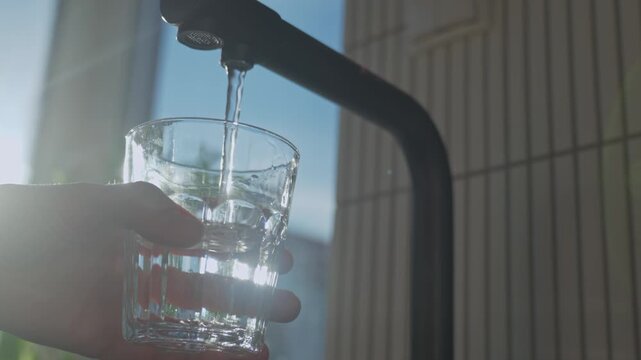 Hand holding glass under black kitchen faucet with sunlight from window in modern home interior. Clean water flowing from tap filter into glass. Concept of drinking water and quenching thirst