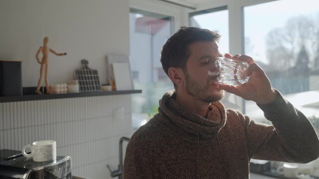 Adult man drinking fresh clean water from glass while standing in kitchen near window with sunlight. Person quenching thirst at home. Concept of hydration and healthy lifestyle