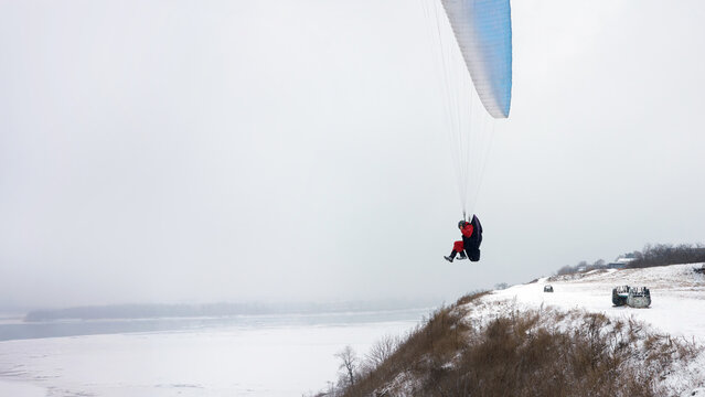 Winter paragliding. A paraglider takes off from a steep, snow-covered river slope. Below, the river is covered in ice.