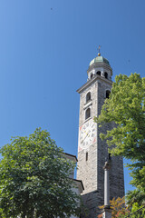 Obraz premium Stone Clock Tower With Green Dome And Cross Under Clear Blue Sky Surrounded By Trees, Lugano Cathedral, Cattedrale di San Lorenzo, Lugano, Switzerland