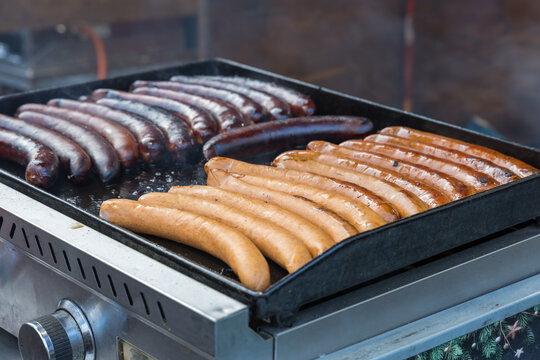 A detailed shot of a large metal griddle flat top grill featuring two rows of sausages of different varieties being cooked. The back row contains darker, smokier sausages