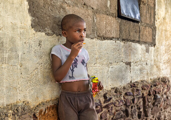 hungry single hungry african girl child in the village, bald shaved head, eating snacks, home next to the house wall,