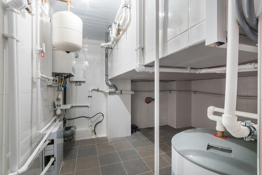 A basement area features a water heater and multiple pipes arranged along the walls. The floor is tiled, and the room has clean lines. Light enters from above.