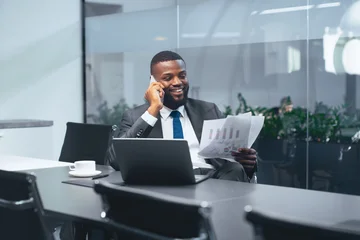 Fotobehang Sportwinkel A businessman is sitting at a table in an office meeting room. He is using a phone while looking at printed documents. A laptop and a cup are on the table with him.  © Prostock-studio