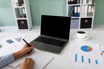 A woman CEO or financial manager participates in an online conference. She takes notes on important information while using her laptop with a blank screen. A coffee cup is nearby.