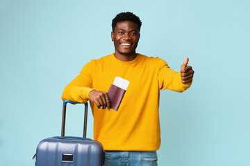 Happy black man with plane tickets, passport and suitcase posing over blue studio background, showing thumb up, copy space. Stylish african american guy travelling while COVID-19 pandemic