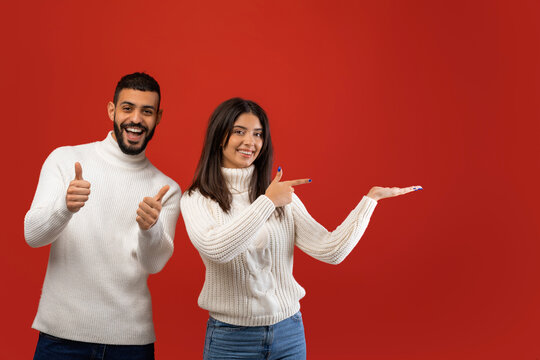 A smiling Arab woman points to an empty space while a man gives a thumbs up, both showcasing excitement for New Year gift ideas against a bold red backdrop in a studio setting.