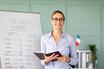 Smiling young woman with glasses stands by a whiteboard featuring French grammar rules. She holds a...