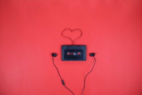 A vintage cassette tape sits on a red surface, with earbud cords forming a heart shape above it. A playful, nostalgic scene that conveys music, love, and creative connection.