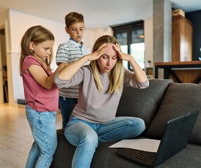 Portrait of a business woman and a mother trying to work on a laptop when her little daughter and son are playing, fooling around and interfering with her. Freelance, work from home, business office
