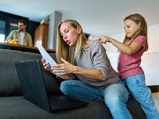 Portrait of a  business woman and a mother trying to work on a laptop when her little daughter is playing, fooling around and interfering with her. Freelance, work from home, business office