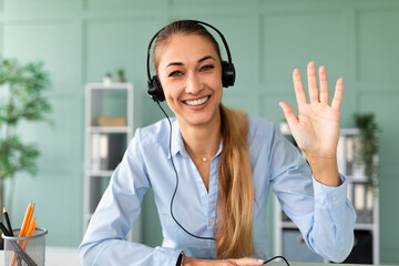 A smiling woman wearing headphones waves at her computer webcam, enjoying a virtual meeting. She sits at her desk, surrounded by a bright and organized workspace, radiating positivity.