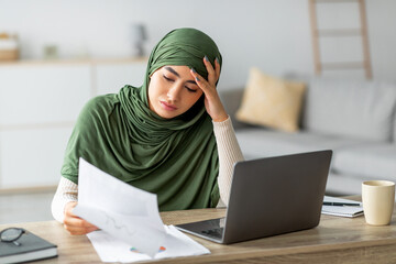 Focused young Arab woman reviews documents while participating in an online business meeting at her home office. She exudes determination as she navigates her work tasks.