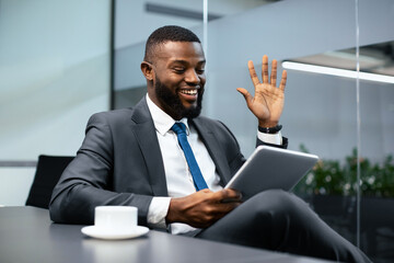 A man in a suit sits at a table in an office, smiling and waving while using a tablet. He appears engaged in a video call. A cup is placed beside him as he communicates during work hours.