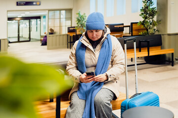 Digital pause and modern solitude emerge as a middle aged woman sits with her suitcase, checking her phone while waiting, surrounded by soft light and a calm travel atmosphere.