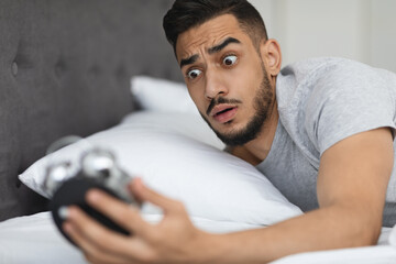 A worried Arab man in bed checks his alarm clock with a shocked expression, realizing he overslept...