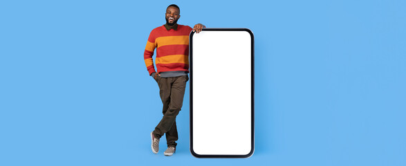 A man wearing a colorful striped sweater stands next to a large blank smartphone against a bright blue background. He appears relaxed and approachable, with a smile on his face.