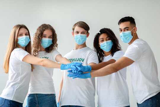 Young diverse volunteers in masks and gloves unite by joining hands, showing teamwork and commitment to volunteer work. They are ready to make a positive impact in their community.