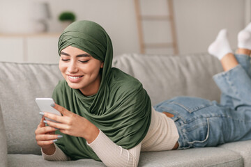 Obraz premium Pretty millennial Arab woman in hijab relaxes on a couch while using her cellphone for work or studies. She enjoys video chatting, immersed in her online tasks at home.