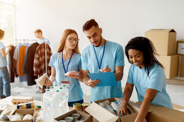 Fototapeta premium Young arab male activist checks a donation list while other volunteers pack boxes with food and supplies. The scene shows teamwork and community support in a charitable organization setting.