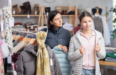Fototapeta premium Adolescence girl try on down jacket in front of mirror during shopping with mother in clothes store. Customer is trying on clothes in store. Buyer is immersed in choice of clothes.
