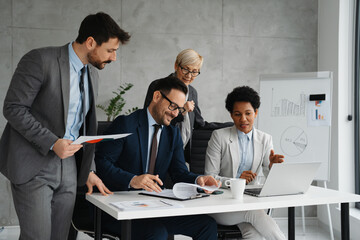 Four diverse business professionals working together at a large desk in a bright, modern office, collaborating on a digital project.