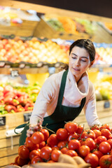 Obraz premium Young woman seller in apron puts tomatoes on display in vegetable shop