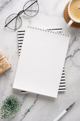 Obraz premium A stylish desk setup featuring a blank notebook on top of striped paper, glasses, a cup of coffee, a small wrapped gift, and seasonal decorations, perfect for holiday planning and creative work