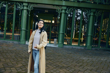 Naklejka premium Woman wearing a long beige coat stands outdoors in front of green glass doors, casual denim and sweater visible, framed by arches and a tiled courtyard beside architectural detail