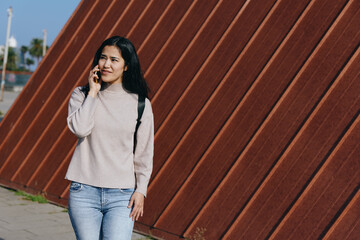 Woman on a sunny outdoor street chats on smartphone, casual sweater and jeans beside a rust colored wall for urban lifestyle, travel vibes and everyday communication
