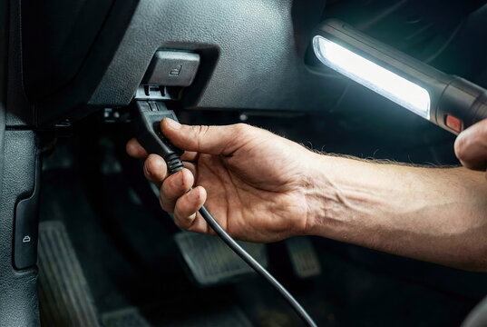 Hand connects car diagnostic scanner to OBD-II port under dashboard, illuminated by flashlight. Essential for vehicle repair and service.