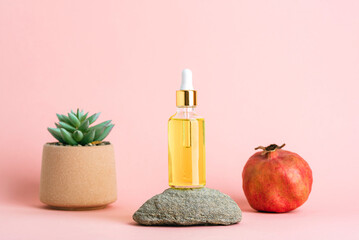 Glass dropper bottle with golden facial oil on stone beside succulent plant and ripe pomegranate on soft pink background