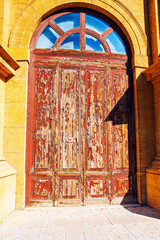 Weathered red wooden door with peeling paint stands within yellow stone archway, topped by segmented transom window. Aged texture and rustic charm evoke history, exposure and architectural character